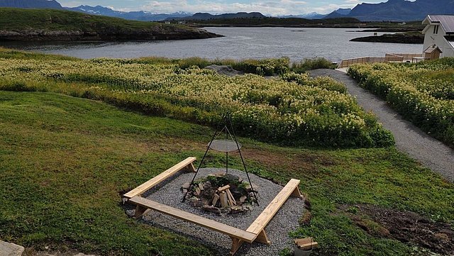 Landschaft in Nordland, Norwegen: Grüne Wiesen, Kiesweg, Feuerstelle mit Sitzgelegenheiten, ruhiger See und malerische Bergkulisse. Perfekt für Erholung und Angelabenteuer in skandinavischer Natur.