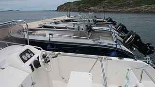 Boote an einem norwegischen Steg in Nord-Norwegen, bereit für Angelabenteuer. Klare Sicht auf Motoren, idealer Ausgangspunkt für Meeresangeln. Friedliche Küstenlandschaft unter bewölktem Himmel lädt zum Entdecken ein.