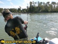 Ein Angler in Tarnkleidung kämpft auf einem Boot mit einer gebogenen Angelrute auf einem glitzernden Fluss, umgeben von grünen Bäumen.