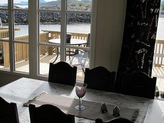 Innenansicht eines Wohnzimmers in Nordland, Norwegen: Blick durch große Fenster auf eine sonnige Holzterrasse mit Tisch am Wasser, umgeben von felsiger Landschaft. Perfekte Kulisse für Angelabenteuer und Entspannung.