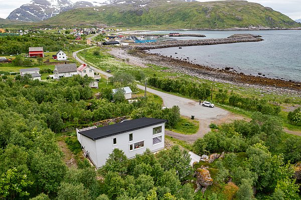 Luftaufnahme einer malerischen Küstenlandschaft in Nord-Norwegen mit weißen Häusern und bewaldeten Hügeln, nahe dem klaren Meer unter schneebedeckten Bergen. Ideal für Naturfreunde und Angelreisen in Norwegen.
