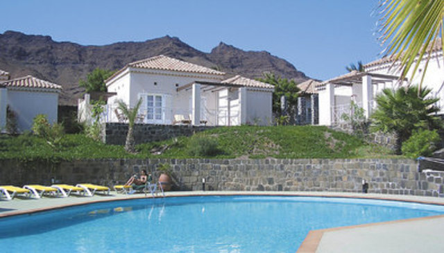 Ein Urlaubsort mit weißen Bungalows und Palmen liegt malerisch vor einer Bergkulisse und verfügt über einen runden Pool mit Sonnenliegen. Ein Urlaubsort mit weißen Bungalows und Palmen liegt malerisch vor einer Bergkulisse und verfügt über einen runden Pool mit Sonnenliegen.