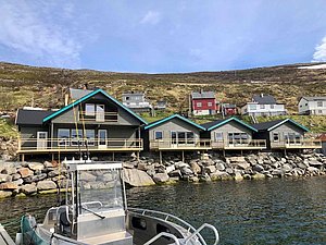 Angelunterkünfte in Nord-Norwegen direkt am Wasser, umgeben von sanften Hügeln. Im Vordergrund ein Boot, ideal für Meeresangeln. Perfekter Ausgangspunkt für einen erholsamen Angelurlaub in malerischer Umgebung.
