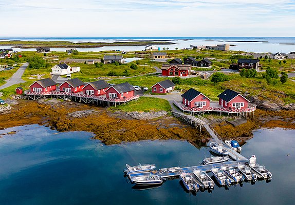 Rote Fischerhütten stehen am Ufer in Nordland, Norwegen, mit Booten vor der Küste. Grüne Landschaft und blauer Himmel schaffen eine idyllische Kulisse für Angelreisen und Naturgenuss im hohen Norden Skandinaviens.