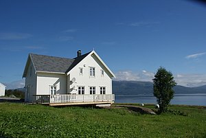 Gemütliches weißes Holzhaus in Nord-Norwegen, umgeben von grüner Wiese und Blick auf den ruhigen Fjord. Ideal für Angelreisen und Naturerlebnisse. Klarer Himmel und Berge im Hintergrund ergänzen die idyllische Szene.