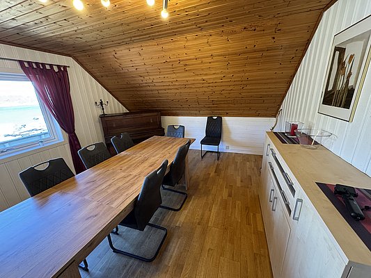 Gemütlicher Meetingraum in einem Dachgeschoss mit Holzwänden und -decke, moderner Holztisch, schwarze Stühle, Klavier, Fenster mit Blick aufs Wasser, rote Vorhänge. Perfekt für Angelurlaube in Hitra, Norwegen.