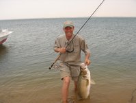 Ein lächelnder Angler steht in flachem Wasser mit einer Angelrute in der Hand und hält einen großen Fisch.