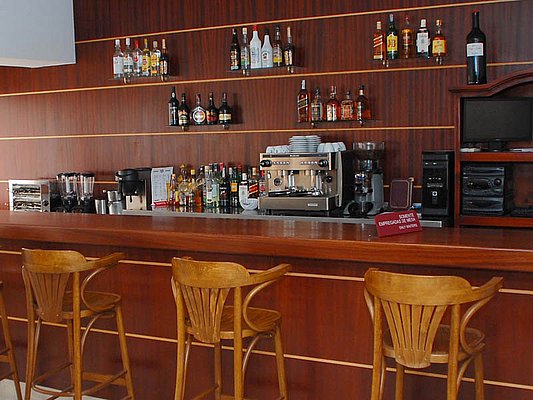 Eine mit Holz verkleidete Bar mit drei Holzstühlen, einer Espressomaschine und einer Auswahl an Spirituosenflaschen auf Regalen. Eine mit Holz verkleidete Bar mit drei Holzstühlen, einer Espressomaschine und einer Auswahl an Spirituosenflaschen auf Regalen.