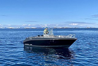 Ein schwarzes Motorboot auf einem ruhigen, blauen Ozean mit schneebedeckten Bergen im Hintergrund.