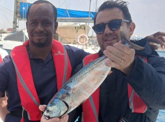 Zwei Personen halten einen frisch gefangenen Fisch vor einem Boot in São Vicente, Kapverden. Sie tragen rote Schwimmwesten. Idealer Angelurlaub für Abenteuerlustige in dieser fischreichen Region.