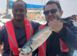 Zwei Personen halten einen frisch gefangenen Fisch vor einem Boot in São Vicente, Kapverden. Sie tragen rote Schwimmwesten. Idealer Angelurlaub für Abenteuerlustige in dieser fischreichen Region.