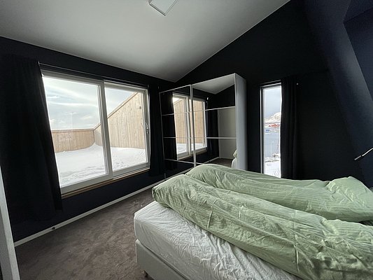 Gemütliches Schlafzimmer in Hitra, Norwegen, mit modernen schwarzen Wänden und komfortabler Bettwäsche. Große Fenster bieten eine atemberaubende Aussicht auf die verschneite Landschaft und das Meer, ideal für einen erholsamen Angelurlaub.