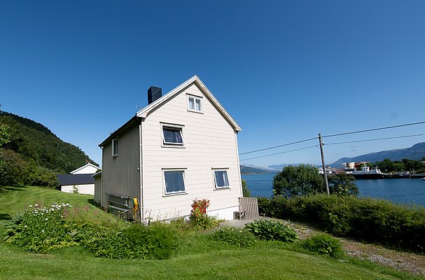 Ein weißes Haus in Nordland, Norwegen, umgeben von saftigem Grün und einem klaren blauen Himmel, mit Blick auf den Fjord und ferne Berge. Ideal für einen entspannten Angelurlaub.