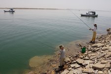 Ein Kind und ein Erwachsener angeln vom felsigen Ufer eines Sees, während zwei Boote im Hintergrund auf dem Wasser treiben.