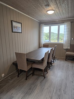 Gemütlicher Raum in Nordland, Norwegen: Holztäfelung, Esstisch mit gepolsterten Stühlen, großes Fenster mit Blick auf grüne Landschaft, gerahmtes Bild an der Wand, skandinavisches Designambiente. Ideal für entspannte Angelreisen mit Komfort.
