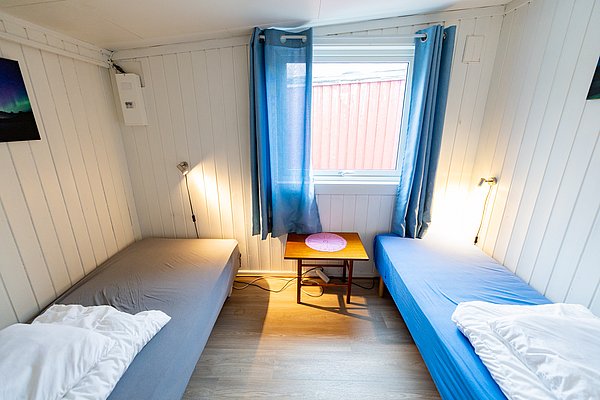 Gemütliches Zimmer in Nordland, Norwegen, mit Holzwänden, zwei Einzelbetten, blauem Vorhang und Fenster. Ideal für Angler auf Angelreise, die nach einer komfortablen Unterkunft suchen. Entspannen nach spannenden Angeltagen.
