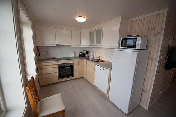 Moderne Küche mit weißer und holzfarbener Einrichtung, ausgestattet mit Kühlschrank, Mikrowelle, Spülmaschine und Herd, sowie einem Holzstuhl am Fenster.