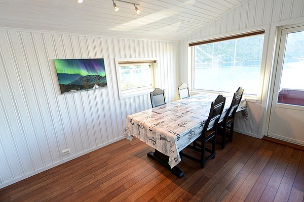 Helles Esszimmer mit Blick auf norwegischen Fjord in Nord-Norwegen, dekoriert mit Polarlicht-Foto. Ideal für entspannten Angelurlaub. Große Fenster und Holzboden schaffen gemütliches Ambiente.