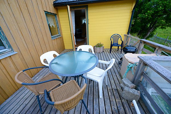 Gemütliche Terrasse eines Holzhauses in Nord-Norwegen, mit Glasstisch und Stühlen, umgeben von grüner Natur. Ideal für entspannte Abende nach aufregenden Angeltouren in dieser idyllischen Umgebung. Perfekte Kulisse für einen Angelurlaub.