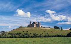 Das Bild zeigt eine mittelalterliche Burg auf einem grünen Hügel unter einem blauen Himmel mit weißen Wolken.
