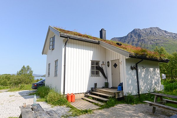 Weißes Landhaus mit begrüntem Dach in Nordland, Norwegen, vor majestätischer Bergkulisse. Ideal für Angelurlaub am Fjord. Nachhaltige Architektur im Einklang mit der Natur. Perfekter Rückzugsort für Outdoor-Abenteuer und Entspannung.