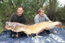 Zwei Männer sitzen im Freien und halten gemeinsam einen großen Wels, der auf einem blauen Tuch liegt.