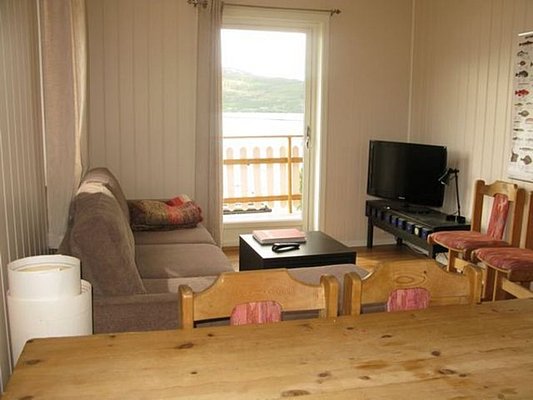Gemütliches Wohnzimmer in Nord-Norwegen mit Sofa, Fernseher und Esstisch. Helle Holzwände und großer Fensterblick auf die malerische Landschaft. Ideal für entspannte Angelurlaub-Entspannung in Skandinavien.