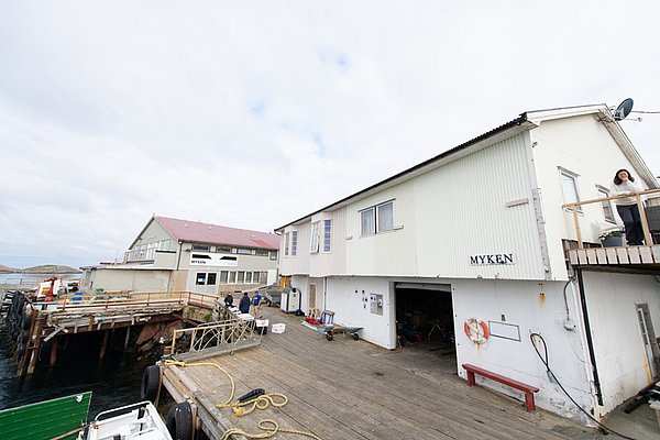 Küstenansicht von Myken, Nordland, Norwegen: Traditionelle Fischereigebäude auf Holzstegen, umgeben von ruhigem Wasser und bewölktem Himmel. Perfekter Ausgangspunkt für Angelreisen in malerischer, friedlicher Umgebung.
