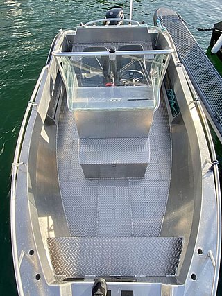 Auf dem Bild ist von oben ein Aluminiumboot zu sehen, das in einem Hafen liegt, mit klar erkennbaren Sitz- und Steuerungseinrichtungen, umgeben von grünem Wasser.