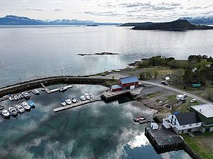 Kleiner Hafen in Nord-Norwegen mit roten Bootshäusern und schneebedeckten Bergen im Hintergrund. Ruhiges, klares Wasser spiegelt die Landschaft. Ideal für Angelurlaub und maritime Abenteuer. Perfekte Mischung aus Natur und Erholung.