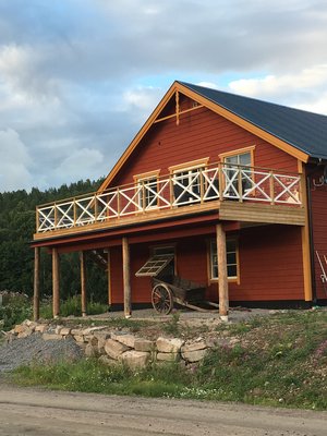 Rotes Holzhaus mit weißem Balkon und Holzwagen in Värmland, Schweden. Ländliche Idylle mit bewölktem Himmel und Wald im Hintergrund – perfekt für einen entspannten Angelurlaub in skandinavischer Natur.