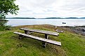 Ein rustikaler Holztisch steht auf einer grünen Wiese mit Blick auf das ruhige Meer und eine kleine Insel. Im Hintergrund liegt ein Boot auf dem Wasser.