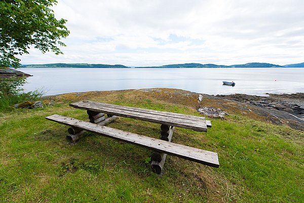 Ein rustikaler Holztisch steht auf einer grünen Wiese mit Blick auf das ruhige Meer und eine kleine Insel. Im Hintergrund liegt ein Boot auf dem Wasser.