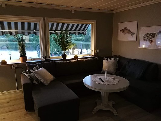 Gemütliches Wohnzimmer mit grauem Ecksofa, rundem Tisch, Pflanzen auf der Fensterbank und Bildern von Fischen an der Wand.