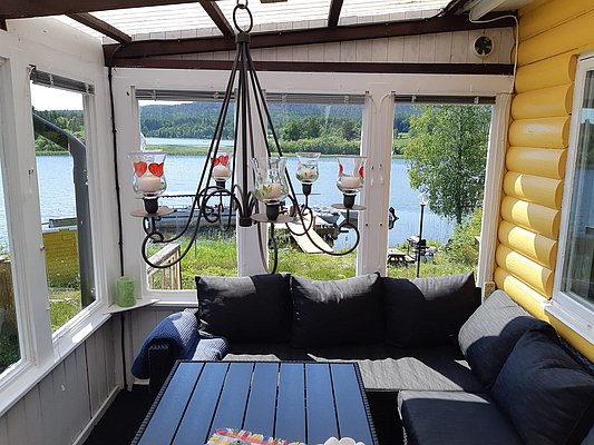 Eine gemütliche Veranda mit schwarzem Sofa und Tisch bietet einen Panoramablick auf einen ruhigen See mit angrenzendem grünen Wald.