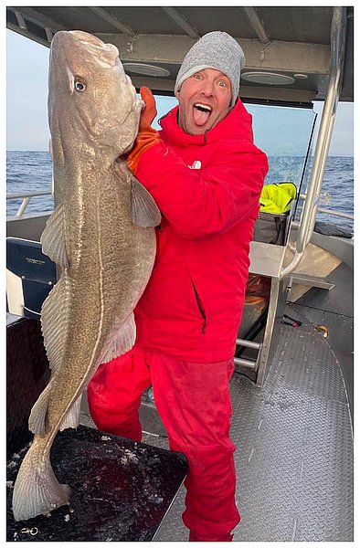 Ein freudiger Angler in roter Kleidung hält einen großen Kabeljau auf einem Boot.