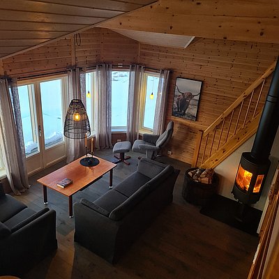 Gemütliches Wohnzimmer in einer Holzhütte in Nord-Norwegen mit Panoramafenstern, Kamin und skandinavischem Design. Perfekt für erholsamen Angelurlaub in der Natur. Ideal zum Entspannen nach einem erfolgreichen Tag am Wasser.