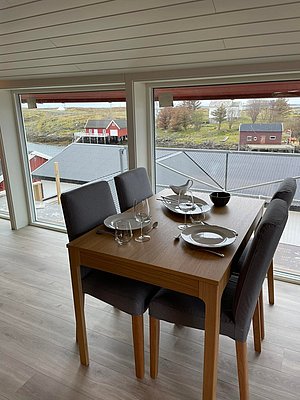 Gemütlicher Essbereich mit gedecktem Tisch, großem Fenster und idyllischem Ausblick auf skandinavische Landschaft mit roten Holzhäusern und Fjord in Nordland, Norwegen. Perfekt für einen erholsamen Angelurlaub.