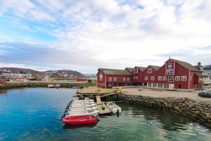 Küste Nord-Norwegens mit roten Gebäuden am Wasser, Booten am Steg und schneebedeckten Hügeln im Hintergrund. Perfekte Kulisse für Angelliebhaber. Ideal für Angelurlaub und Meeresangeln in idyllischer, ruhiger Umgebung.