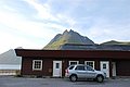 Rotes Holzgebäude vor majestätischen Bergen in Nordland, Norwegen. Ein silberner SUV parkt davor, während der klare Himmel den Blick auf die beeindruckende Landschaft freigibt. Perfekter Ausgangspunkt für Angelreisen in der Region.