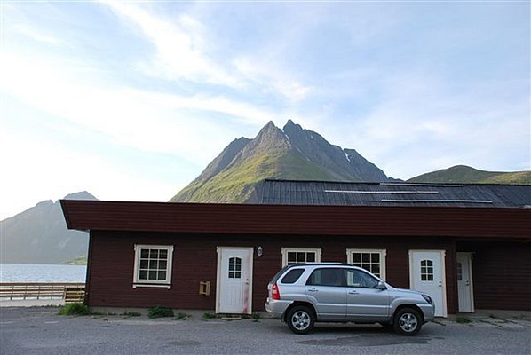 Rotes Holzgebäude vor majestätischen Bergen in Nordland, Norwegen. Ein silberner SUV parkt davor, während der klare Himmel den Blick auf die beeindruckende Landschaft freigibt. Perfekter Ausgangspunkt für Angelreisen in der Region.