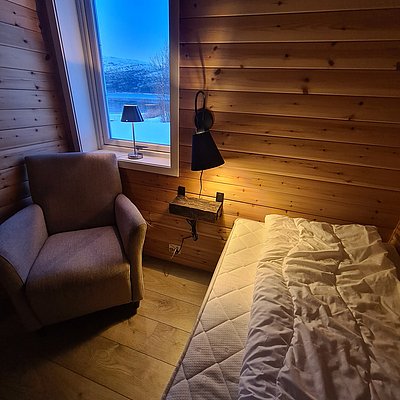Gemütliches Holzzimmer in Nord-Norwegen mit Bett, Leselampe und Sessel. Großes Fenster bietet atemberaubenden Blick auf schneebedeckte Landschaft. Perfekter Rückzugsort nach einem abenteuerlichen Angeltag.