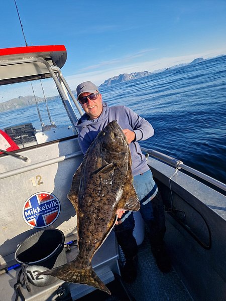 Ein Angler hält stolz einen großen Fisch auf einem Boot, umgeben von der beeindruckenden norwegischen Küstenlandschaft. Klare blaue Himmel und ruhige See verleihen dem Angelabenteuer eine malerische Kulisse.