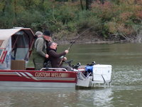 Drei Personen sind auf einem Boot auf einem Fluss beim Angeln zu sehen, wobei einer der Angler eine Angelrute hält, während die anderen zuschauen.