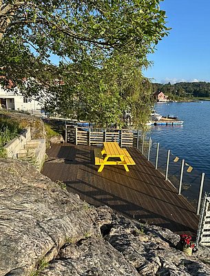 Sonniges Holzdeck in Hitra, Norwegen: Gelbe Picknickbank unter grünem Baum, mit Blick auf einladendes Wasser. Ideal für entspannte Angelpausen. Natur und Ruhe vereint. Perfekter Ort für deinen nächsten Angelurlaub.