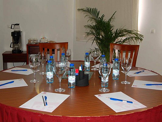 Ein Konferenzraum mit einem runden Tisch, der mit Wasserflaschen, Weingläsern, Notizblöcken und Stiften gedeckt ist, im Hintergrund eine Kaffeestation und eine große Pflanze. Ein Konferenzraum mit einem runden Tisch, der mit Wasserflaschen, Weingläsern, Notizblöcken und Stiften gedeckt ist, im Hintergrund eine Kaffeestation und eine große Pflanze.