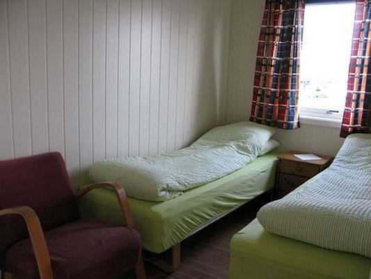 Gemütliches Schlafzimmer in Nordland, Norwegen: Zwei Einzelbetten mit grüner Bettwäsche, ein Holztisch, roter Sessel, helle Holzverkleidung, Fenster mit bunter Gardine. Perfekt für erholsame Angelreisen und Entspannung in skandinavischer Natur.
