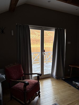 Sonnendurchfluteter Raum in Nord-Norwegen, Blick durch Fenstertüren auf verschneite Landschaft und Fjord. Gemütlicher Holzstuhl im Vordergrund. Perfekte Kulisse für erholsamen Angelurlaub in Norwegen.