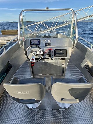 Ein Boot mit zwei Sitzen und einem Steuerstand aus Metall liegt auf einem ruhigen Gewässer mit einer malerischen Aussicht auf die bewaldete Küstenlinie und den klaren blauen Himmel im Hintergrund.