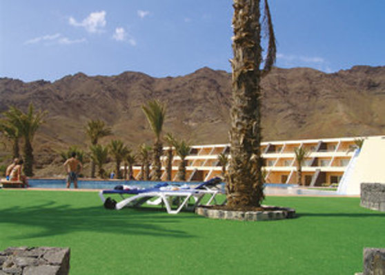 Die Fotografie zeigt einen Poolbereich mit Liegen, umgeben von Palmen, vor einer bergigen Kulisse und einem mehrstöckigen Gebäude im Hintergrund. Die Fotografie zeigt einen Poolbereich mit Liegen, umgeben von Palmen, vor einer bergigen Kulisse und einem mehrstöckigen Gebäude im Hintergrund.